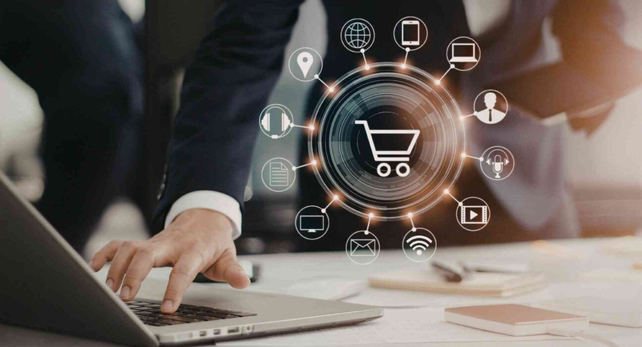Osoba pracująca na laptopie z graficznym symbolem koszyka i ikonami kanałów digital wokół – koncepcja rozwoju e-commerce i sprzedaży wielokanałowej.