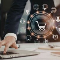 Osoba pracująca na laptopie z graficznym symbolem koszyka i ikonami kanałów digital wokół – koncepcja rozwoju e-commerce i sprzedaży wielokanałowej.