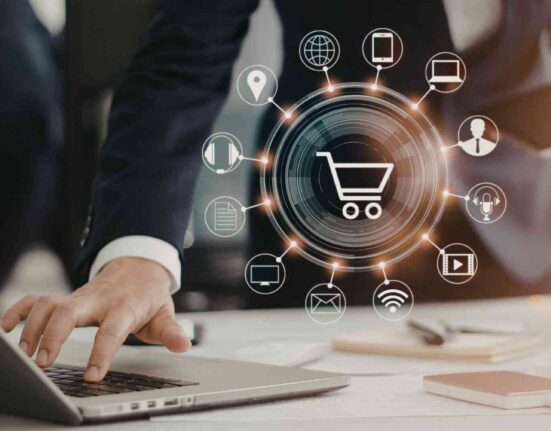 Osoba pracująca na laptopie z graficznym symbolem koszyka i ikonami kanałów digital wokół – koncepcja rozwoju e-commerce i sprzedaży wielokanałowej.